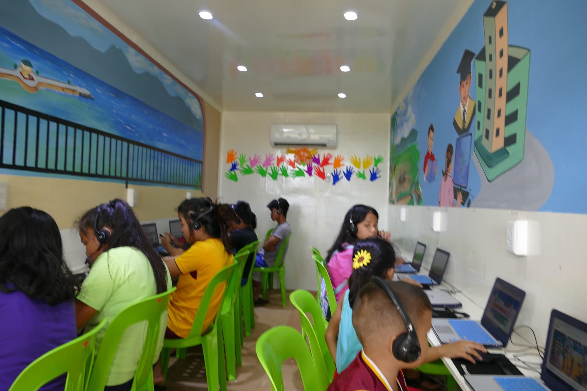 A full class at laptops inside the container classroom, murals on the walls
